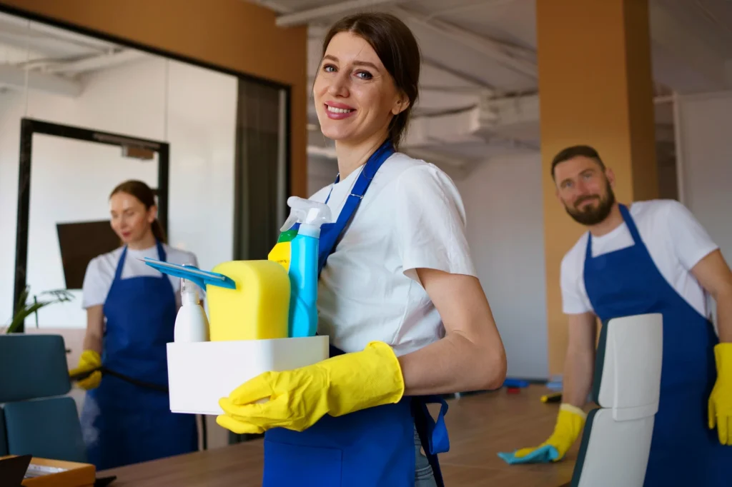 Smiling professional cleaner holding cleaning supplies with team in office environment