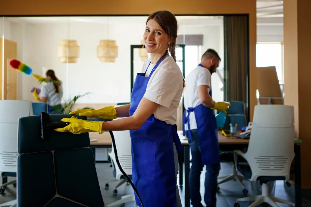 Female cleaner in blue apron and yellow gloves using a floor buffer in a commercial office space.